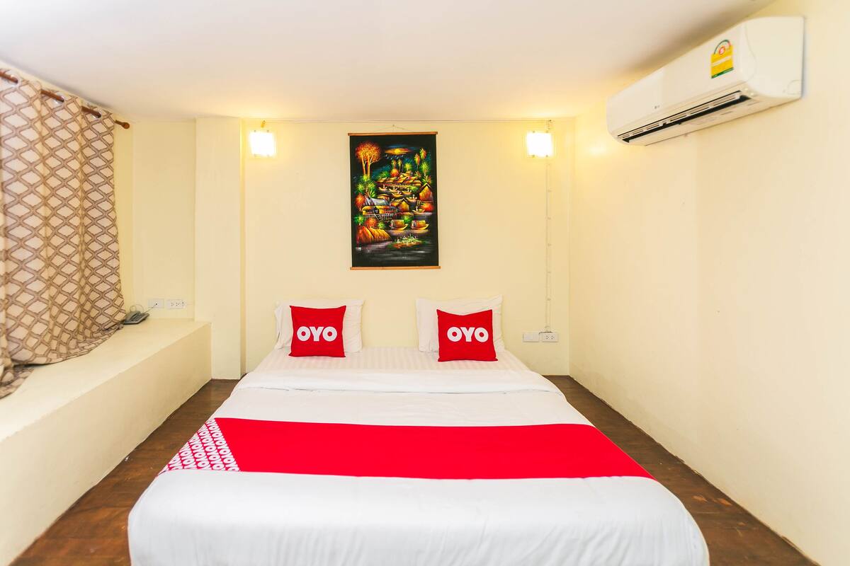 A hotel room with a bed, two pillows with "OYO" branding, a wall-mounted air conditioner, and a painting above the bed.