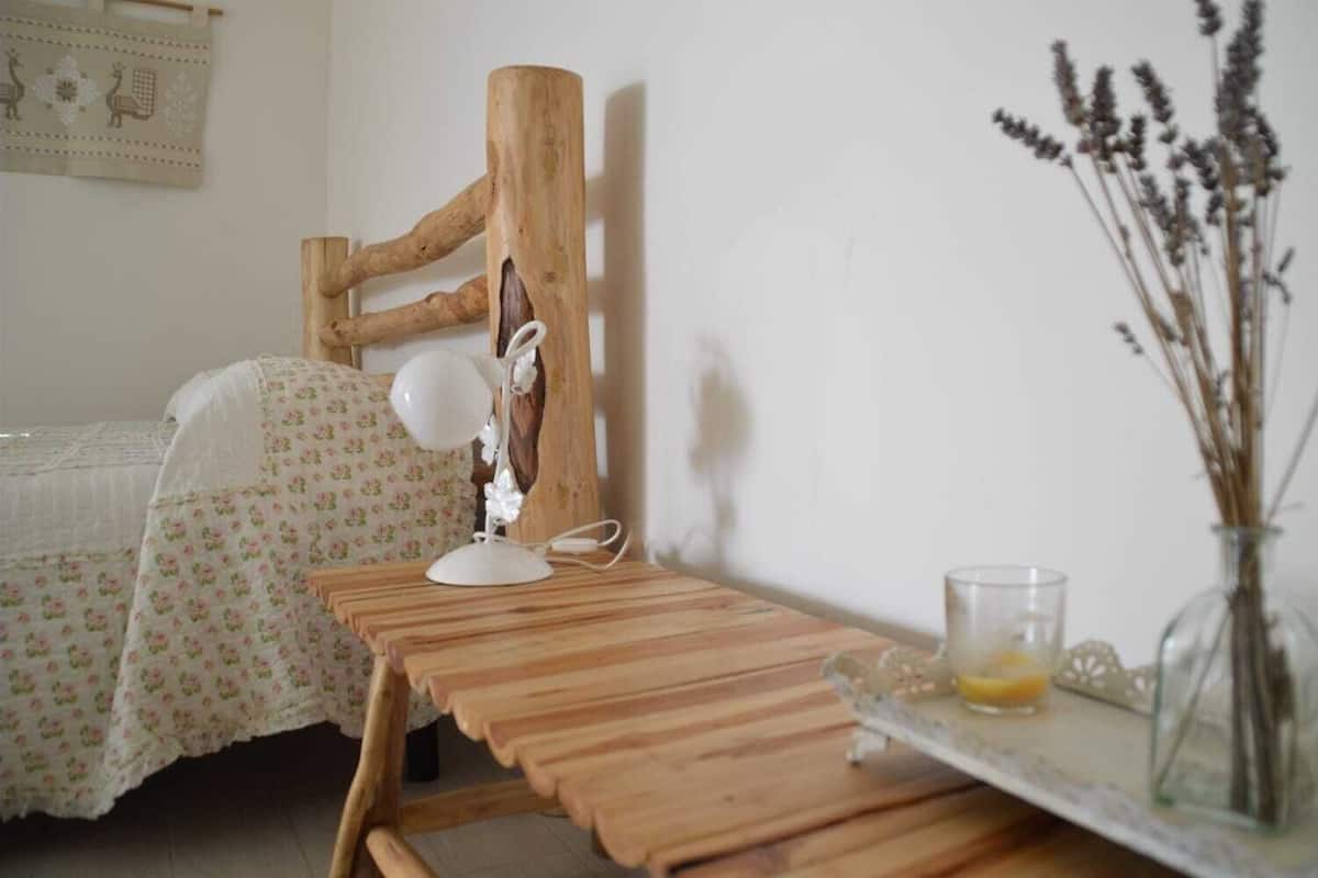 Uma mesa de madeira com uma luminária, um vaso de vidro com plantas secas e uma cama com roupa de cama florida.