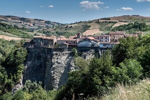 A village with buildings perched on a cliff edge, surrounded by greenery and hills. - Il Pavone Errante (Mercato Saraceno)