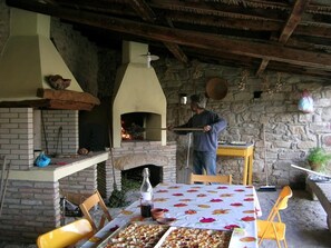 A rustic outdoor dining area with a brick fireplace, a table set with food, and a person cooking. - Sa Domu De Sa Contissa (Selegas)