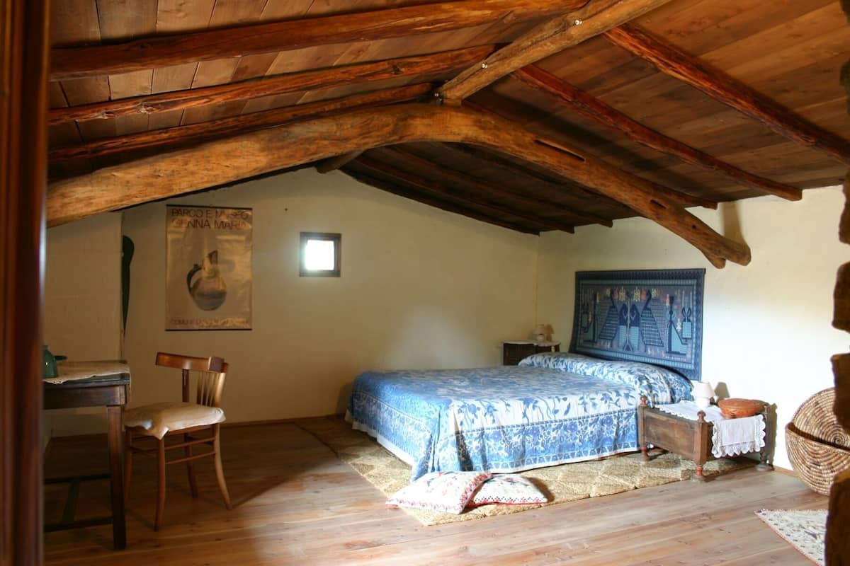 Ein rustikales Schlafzimmer mit Holzbalkendecke, einem Bett mit gemustertem Überwurf, einem Stuhl, einem kleinen Tisch und einer wandhängenden Wandteppich.