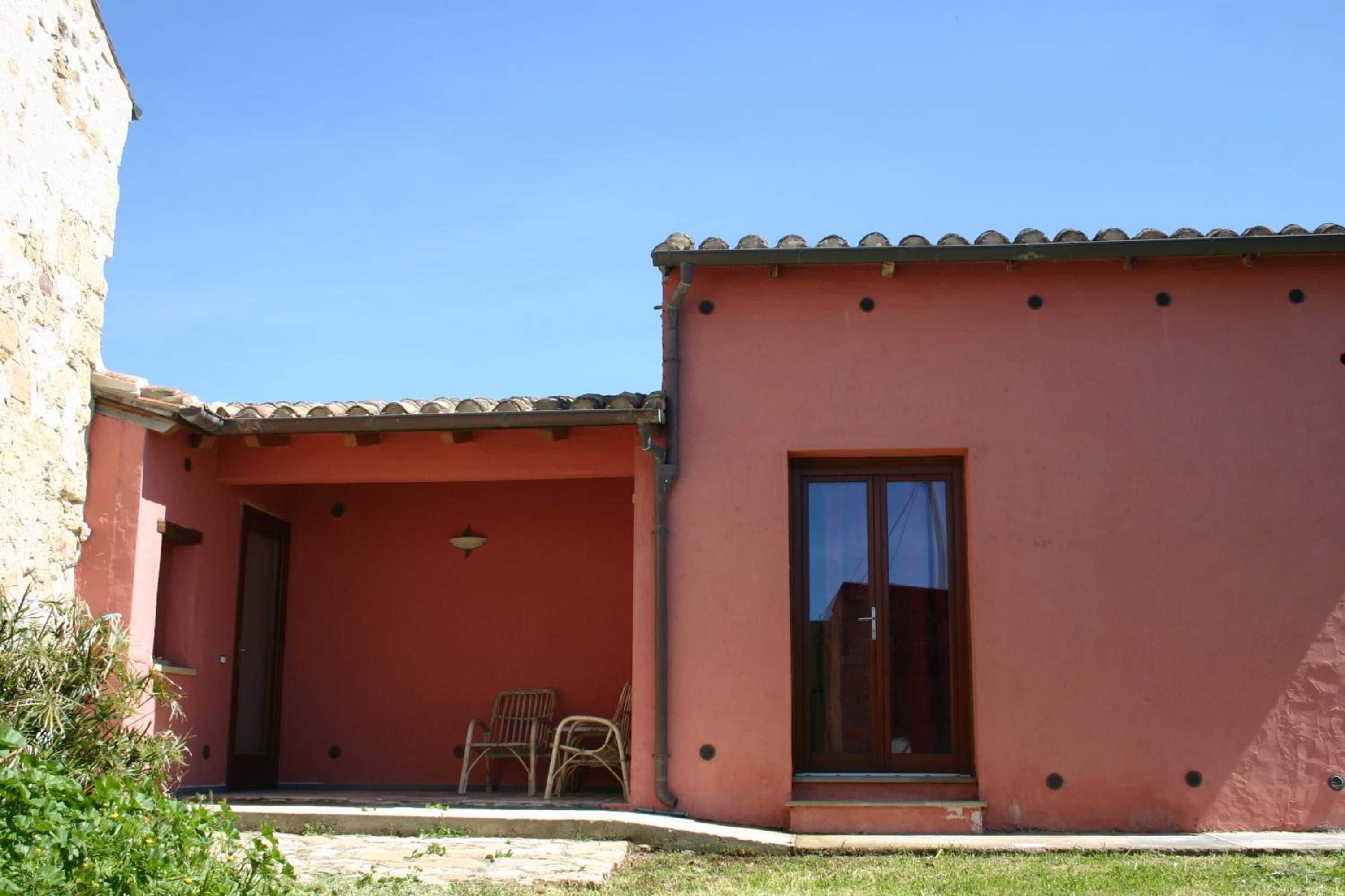 Un bâtiment à l’extérieur rouge, avec un toit en tuiles et une porte en verre. On peut voir deux chaises en osier à l’extérieur.