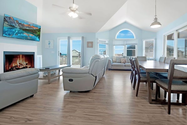 A spacious living room with a fireplace, a large TV, a reclining sofa, a dining area with wooden chairs, and large windows offering a view of the outside.