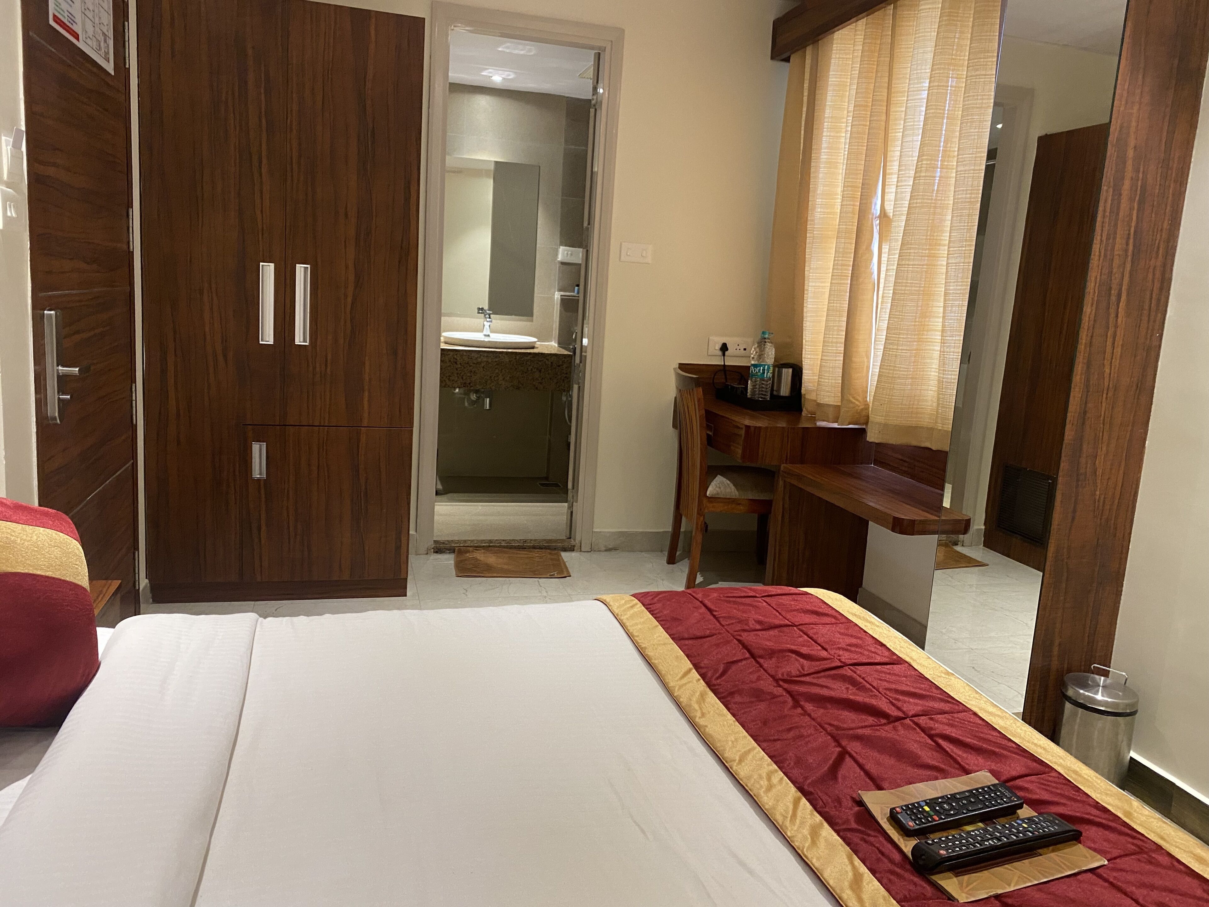 A hotel room with a bed, a wooden wardrobe, a desk with a chair, a bathroom with a sink and mirror, and a trash bin.