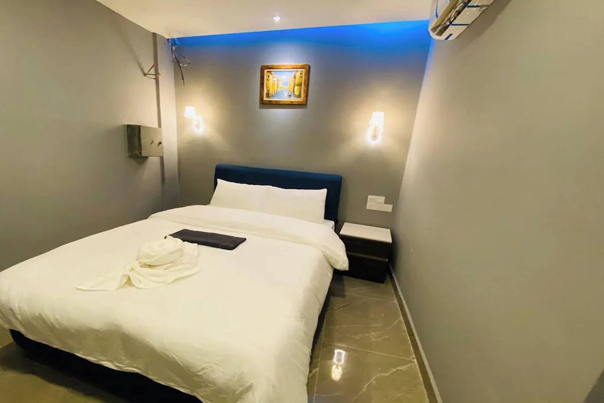 Una cama bien tendida con sábanas blancas y una toalla, una mesita de noche y un aplique de pared.