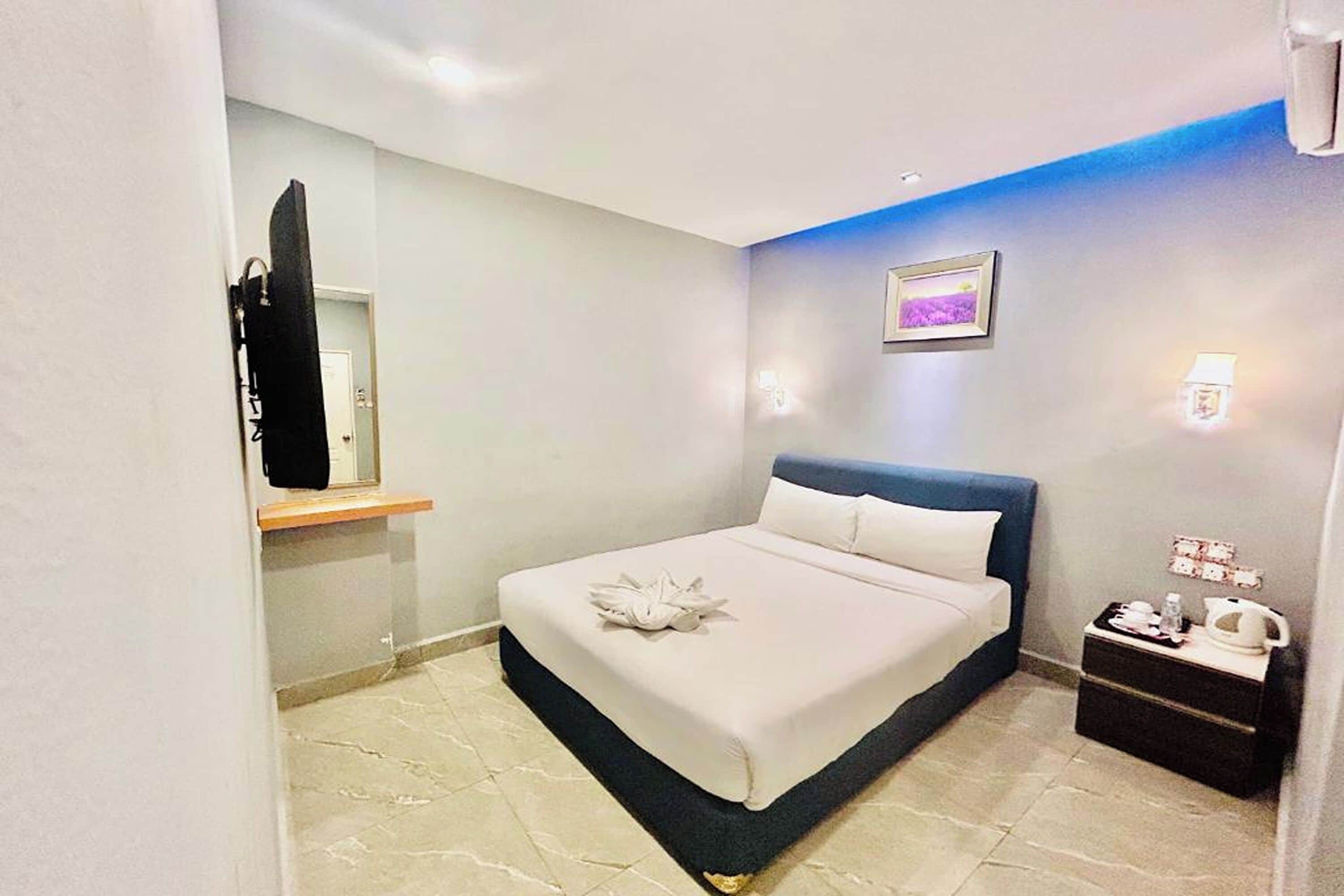 A hotel room with a bed, a nightstand, a mirror, a picture on the wall, and a wall-mounted light.