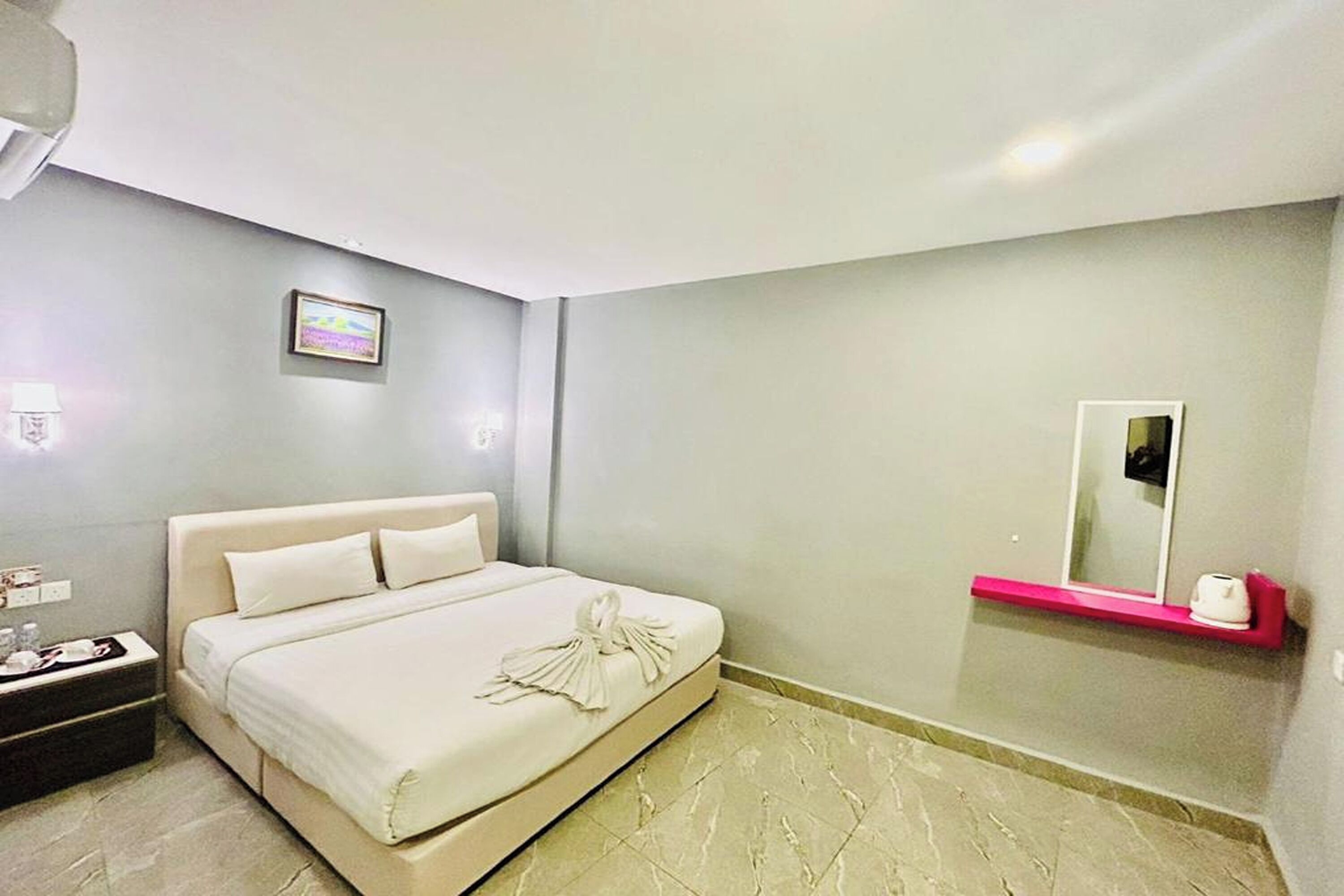 A modern hotel room with a large bed, a bedside table with a lamp, a small shelf with a mirror, and a picture on the wall.