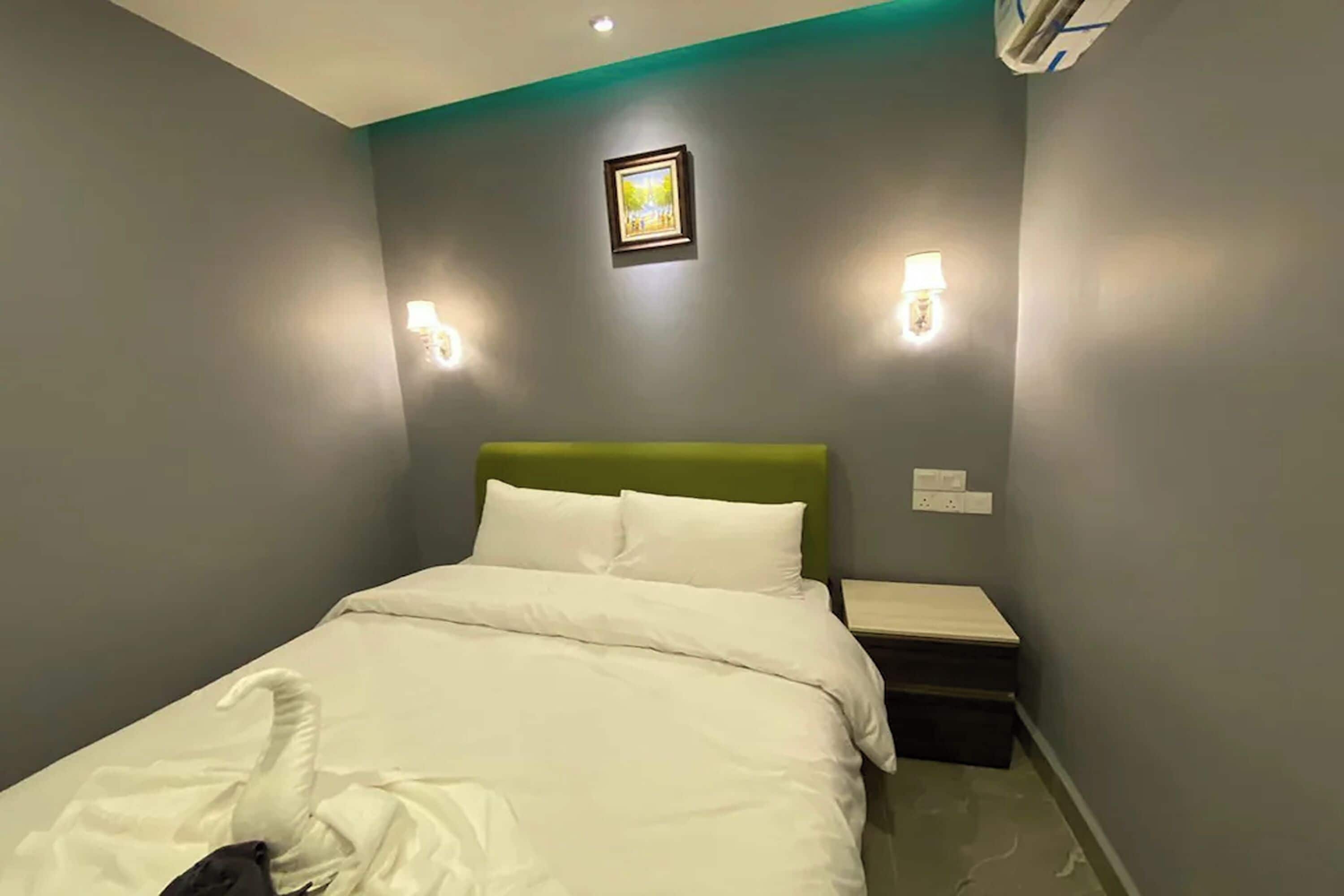 Una cama bien hecha con ropa de cama blanca, un cabecero verde y dos mesitas de noche fijadas a la pared.