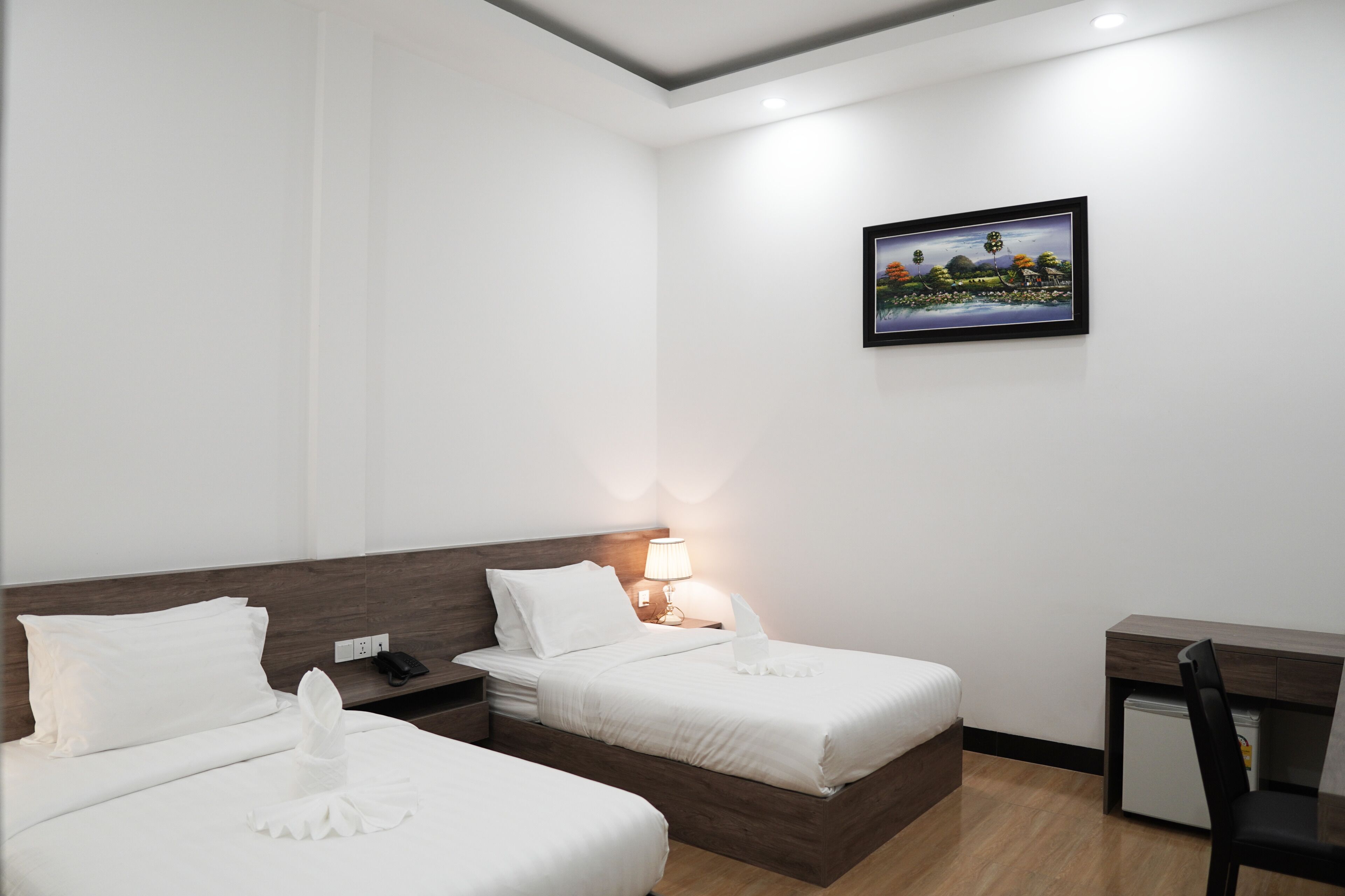 A hotel room with two beds, a desk, a chair, a lamp, and a painting on the wall.