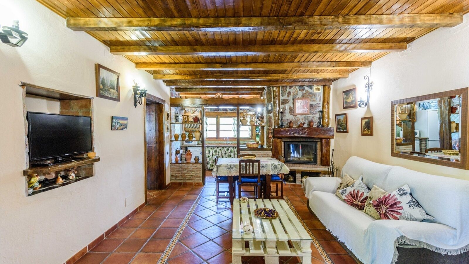 A cozy living room with a wooden ceiling, a TV mounted on the wall, a fireplace, and a sofa with floral cushions.