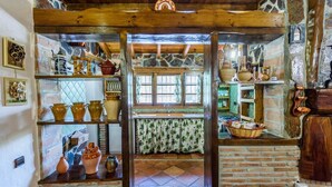Uma cozinha rústica com paredes de pedra, vigas de madeira e prateleiras repletas de cerâmica.