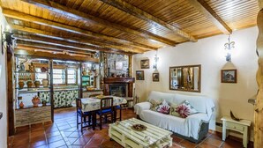 Interior rústico com teto de madeira, sofá branco, mesa de jantar com cadeiras e cozinha com lareira.
