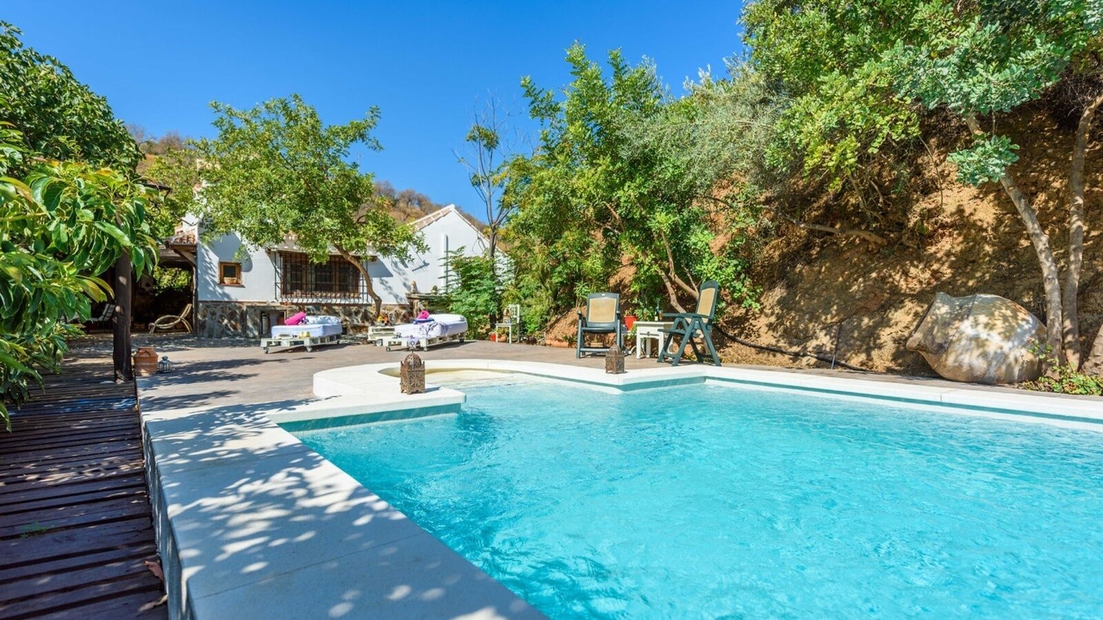 A swimming pool with a wooden deck, lounge chairs, and a white building in the background.