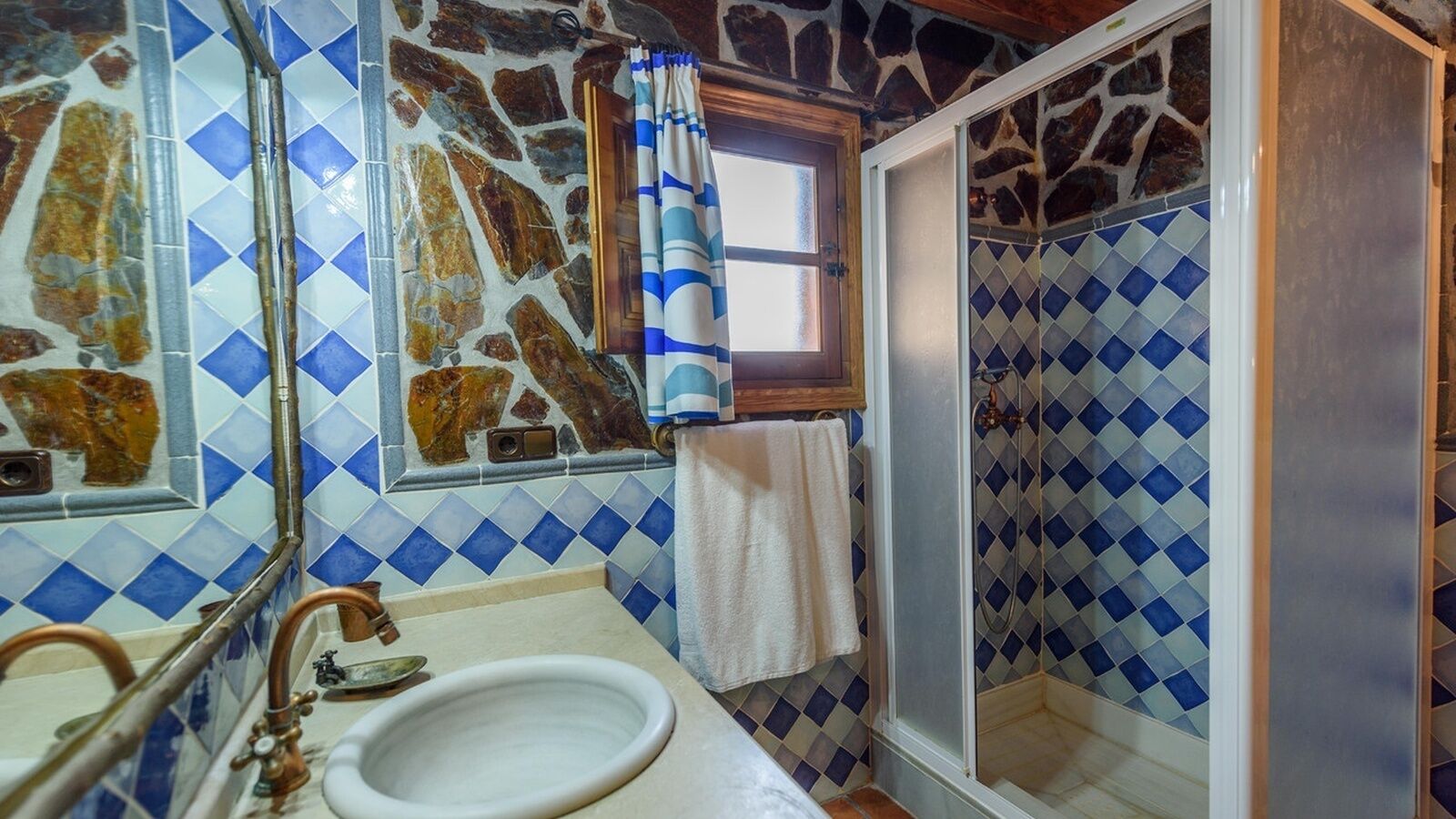 A bathroom with a stone mosaic wall, a white sink, and a shower with a blue and white tile pattern.