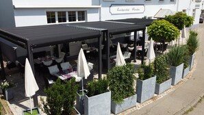 An outdoor dining area with tables and umbrellas, surrounded by planters and a building with a sign. - Landgasthof Hirschen (Hohentengen am Hochrhein)
