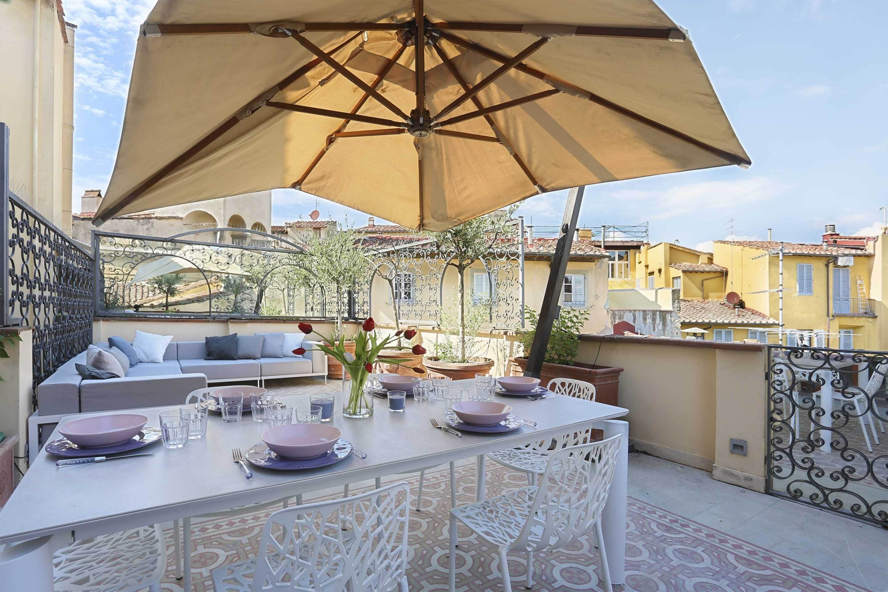 Une terrasse avec une table à manger pour quatre personnes, entourée de chaises blanches, sous un grand parasol.