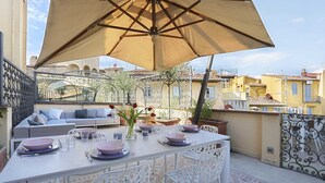 A terrace with a dining table set for four, surrounded by white chairs, under a large umbrella.