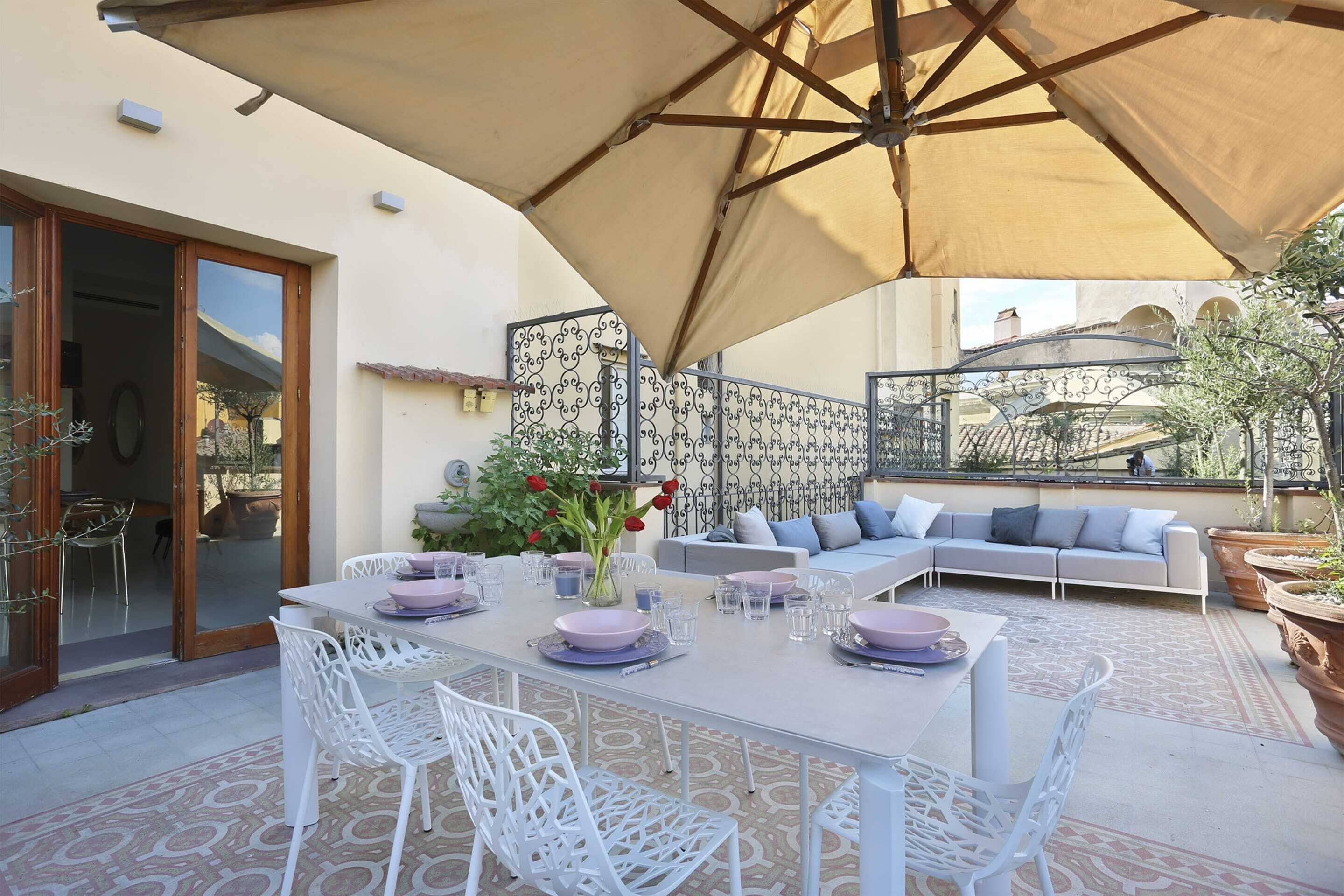 Un espace de restauration en plein air avec une table dressée pour quatre personnes, entourée de chaises blanches, sous un grand parasol.