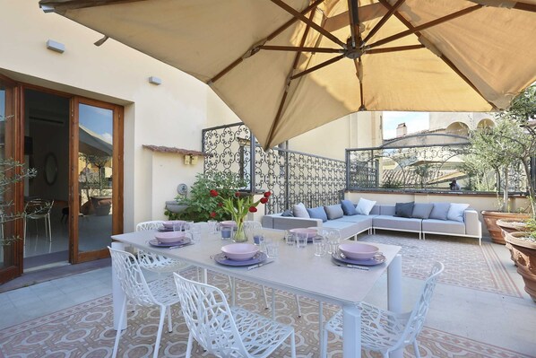 Un espace de restauration en plein air avec une table dressée pour quatre personnes, entourée de chaises blanches, sous un grand parasol.