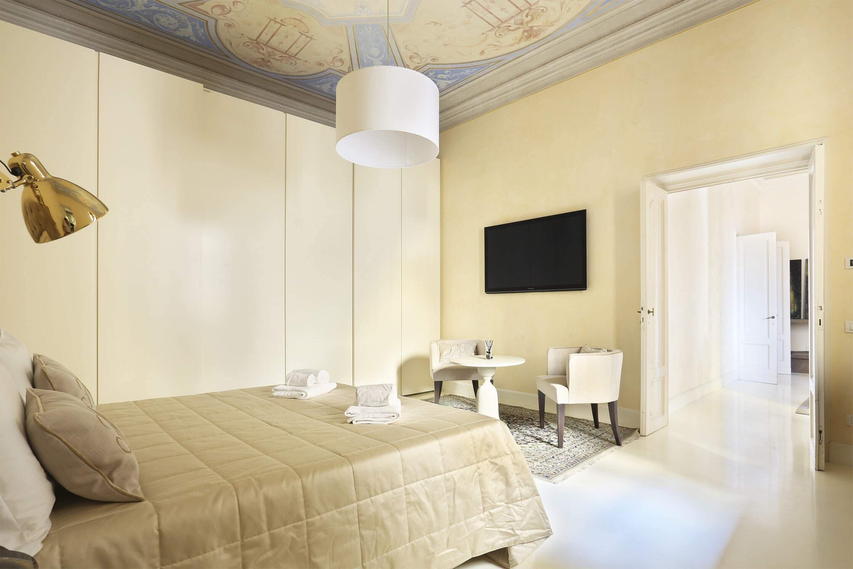 Ein Hotelzimmer mit einem Bett, einem Schreibtisch, zwei Stühlen, einem Fernseher und einer Tür zu einem weiteren Raum.