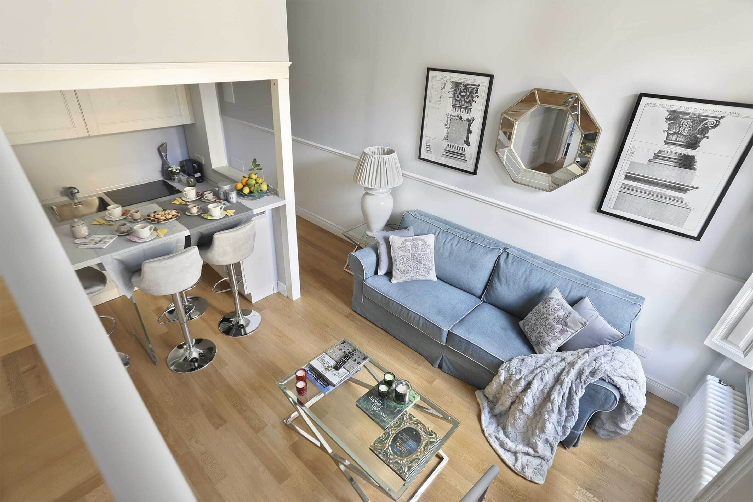 Ein modernes Wohnzimmer mit einem blauen Sofa, einem Glastisch und einer Küchenzeile inklusive Esstisch und Barhocker.