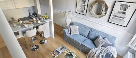 Ein modernes Wohnzimmer mit einem blauen Sofa, einem Glastisch und einer Küchenzeile inklusive Esstisch und Barhocker.