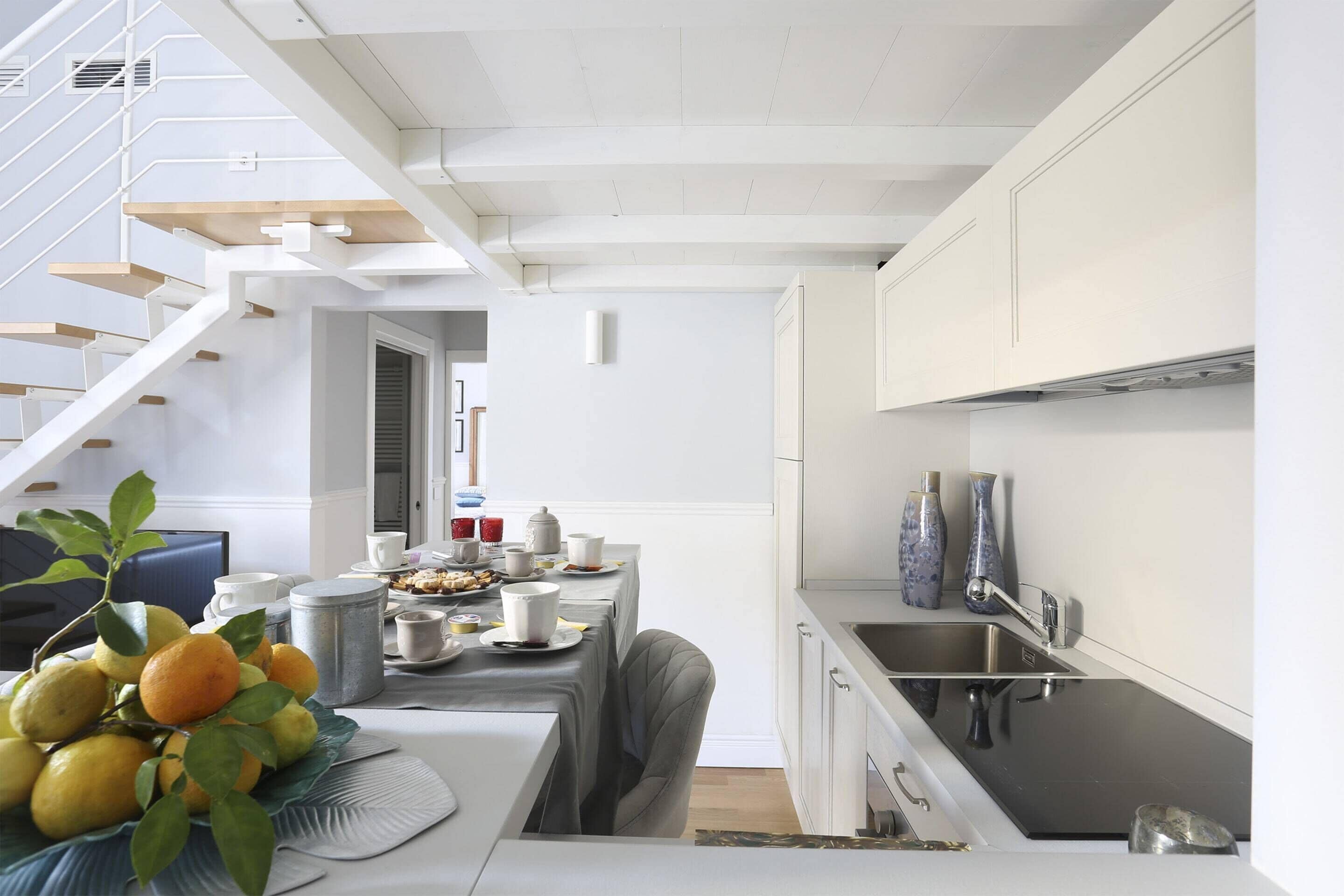 Cozinha moderna com armários brancos, uma pia de aço inoxidável e um espaço de refeições com mesa posta para dois.
