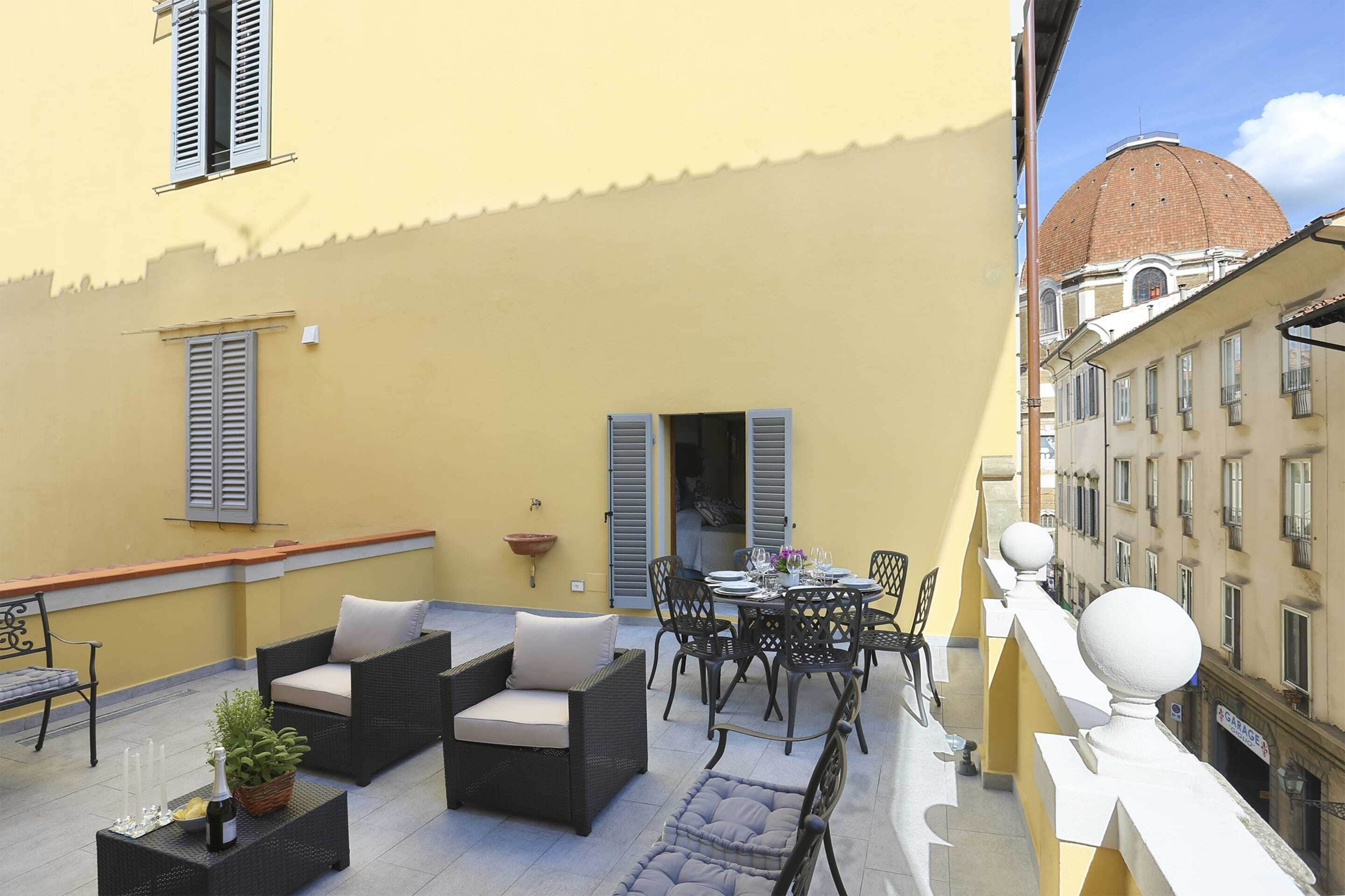 An outdoor patio with wicker furniture, a dining table set for four, and a view of a building with a dome.