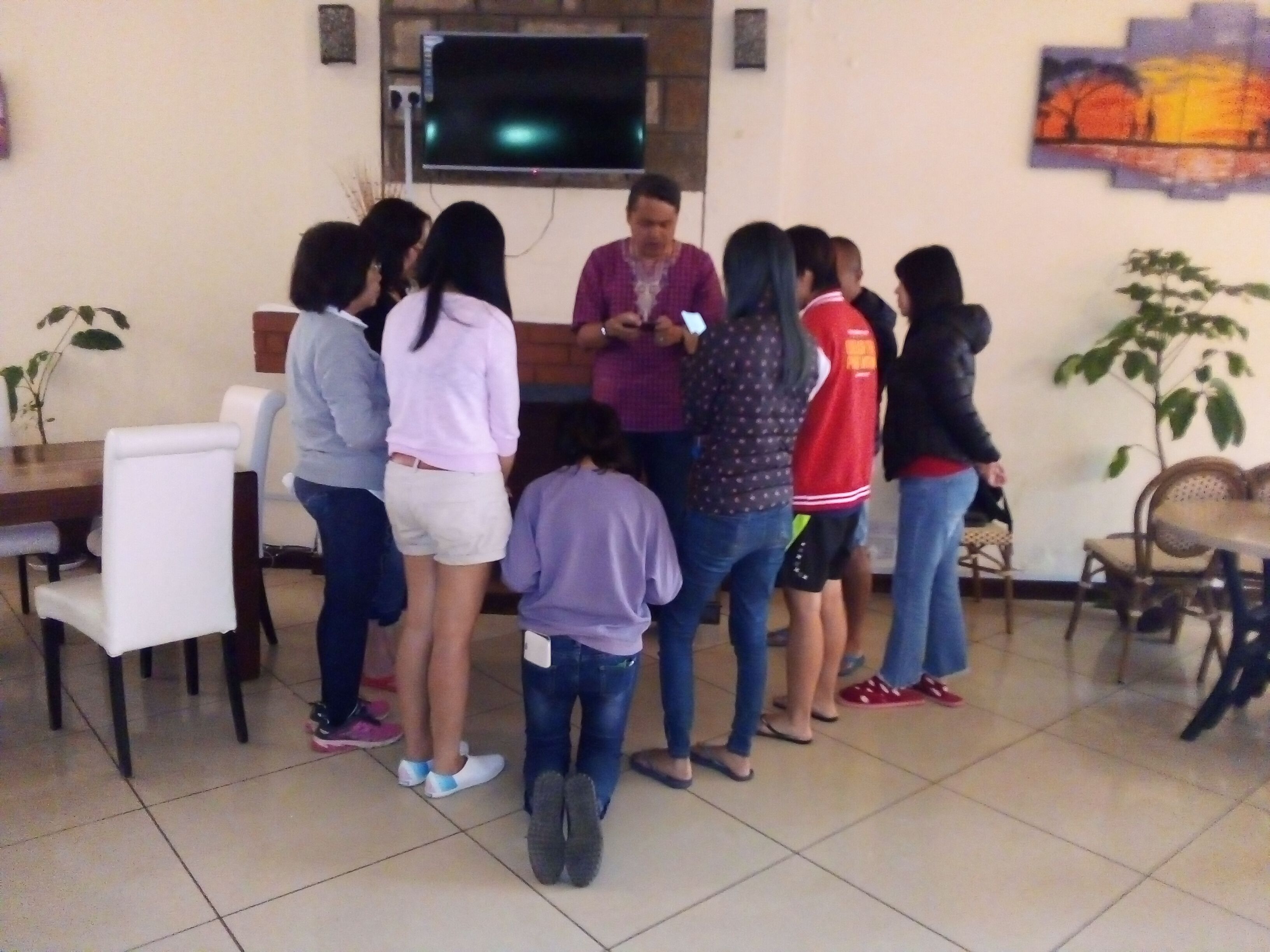 A group of people in a room with a television mounted on the wall, a dining table, and chairs.