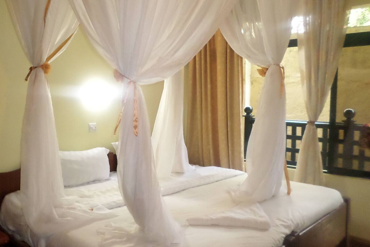 A room with a bed under a canopy, a nightstand, a wall socket, and a window with curtains.