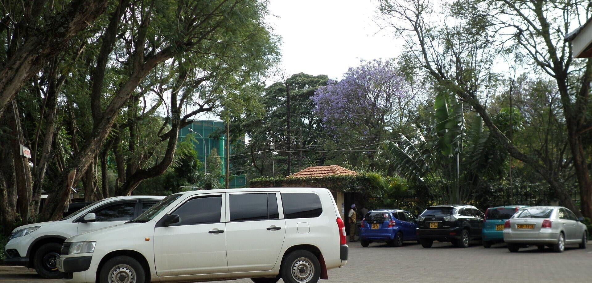 A parking area with several cars, including a white van, parked among trees and a building with a tiled roof.