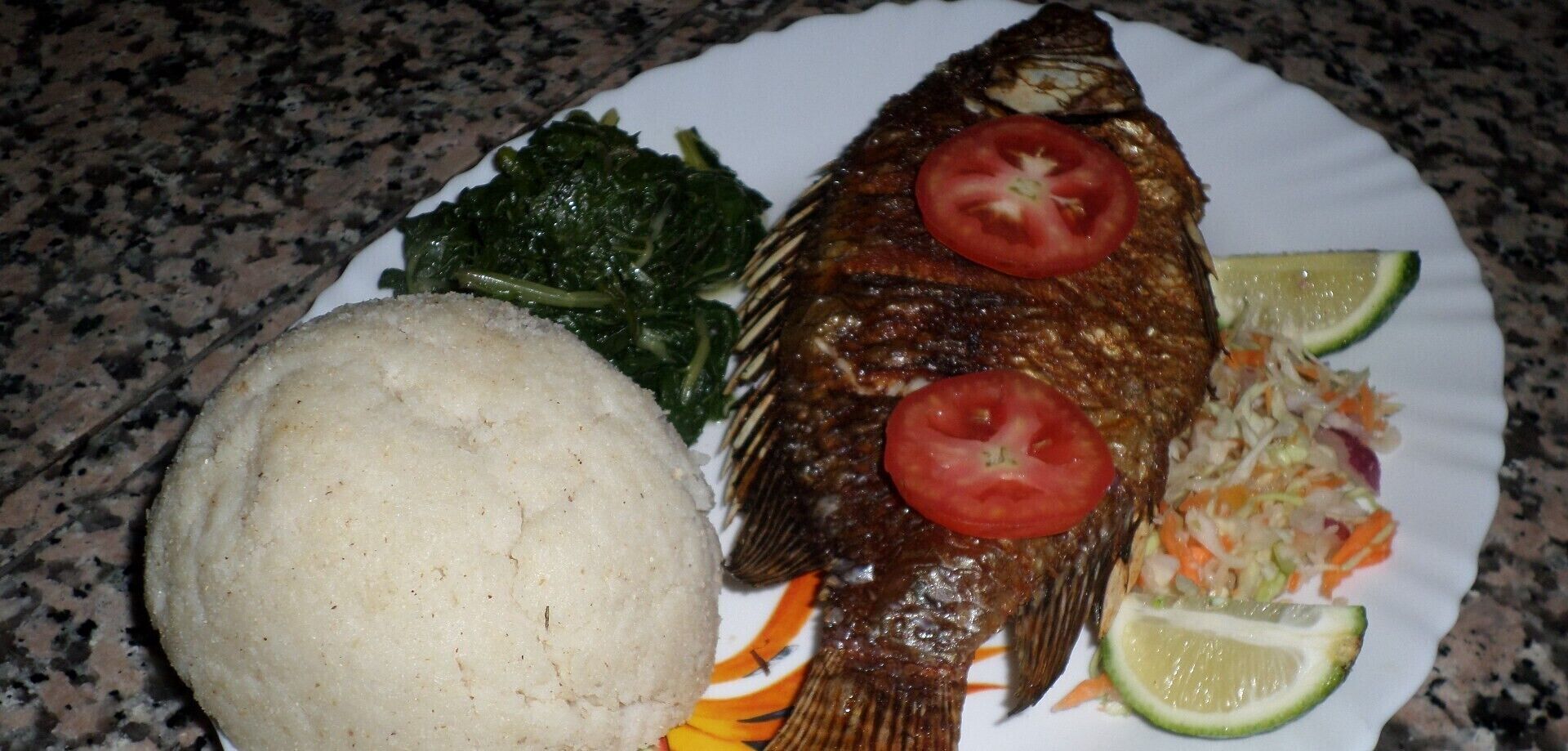 A plate with a whole fish, two slices of tomato, a slice of lime, a serving of greens, and a mound of what appears to be a grain-based dish.