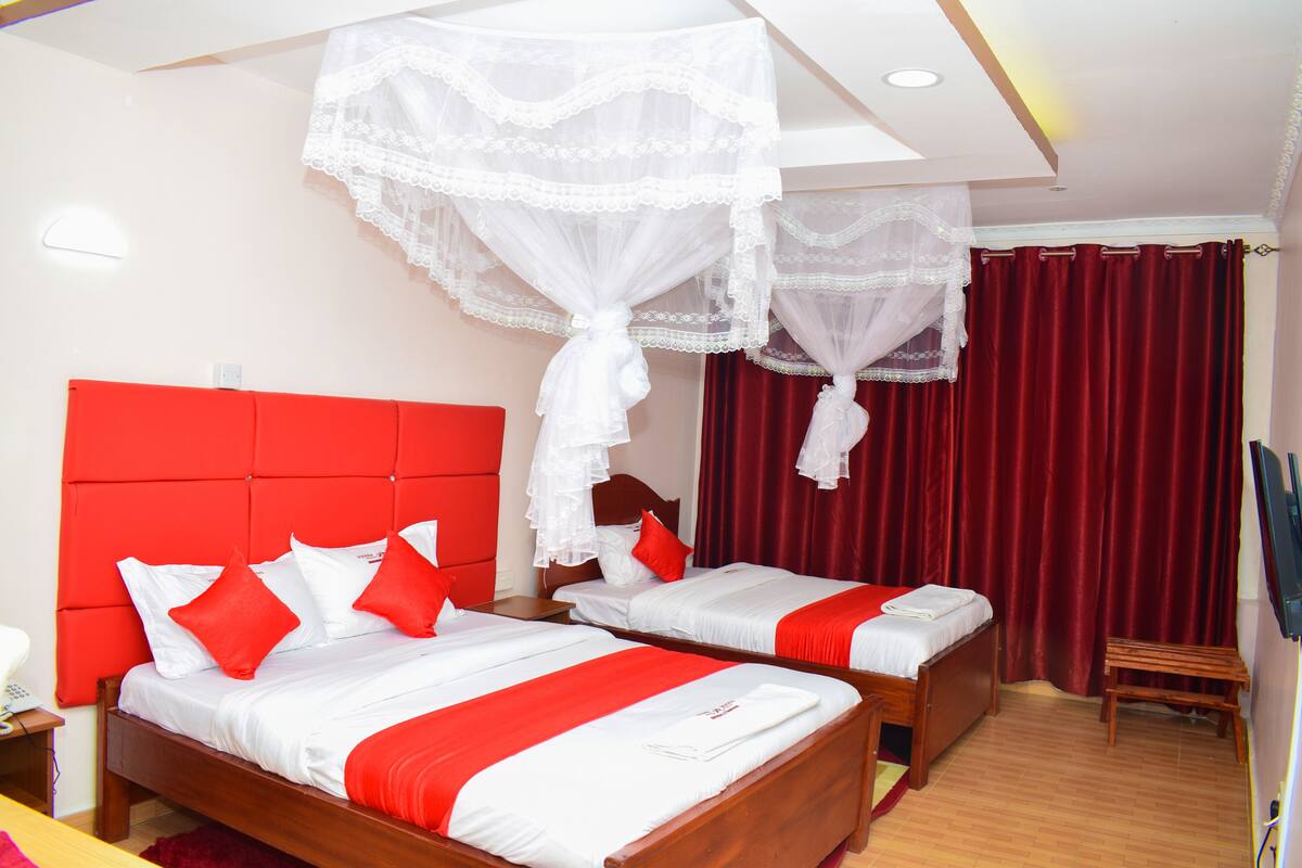 A hotel room with two beds, red and white bedding, a red headboard, and a wooden bench.