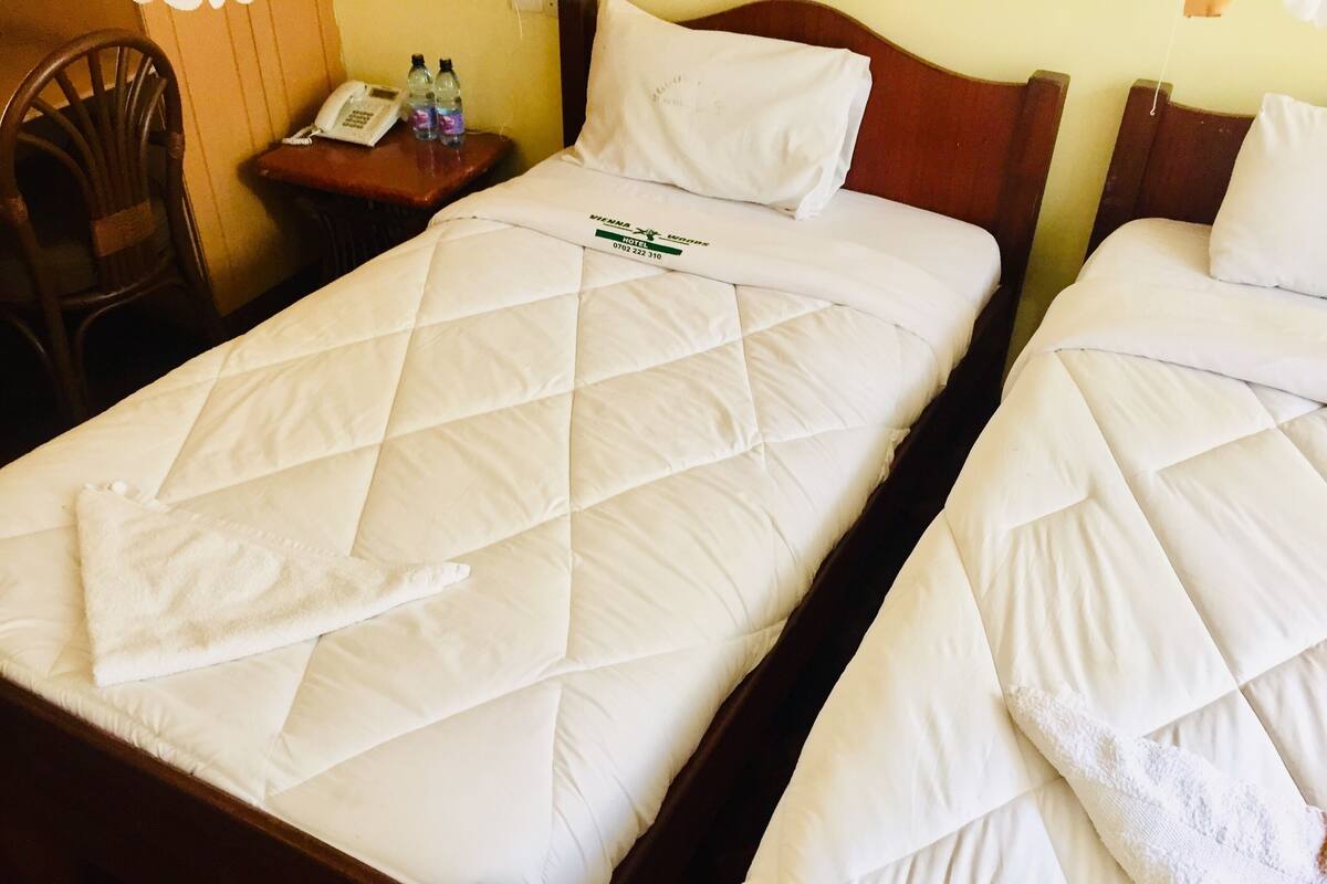 A hotel room with two beds, each with a quilted white duvet and pillows. There is a wooden nightstand with a telephone and bottled water.