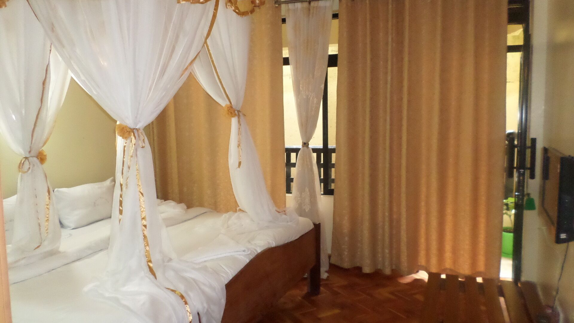 A four-poster bed with white linens and gold accents, a wooden floor, and beige curtains.