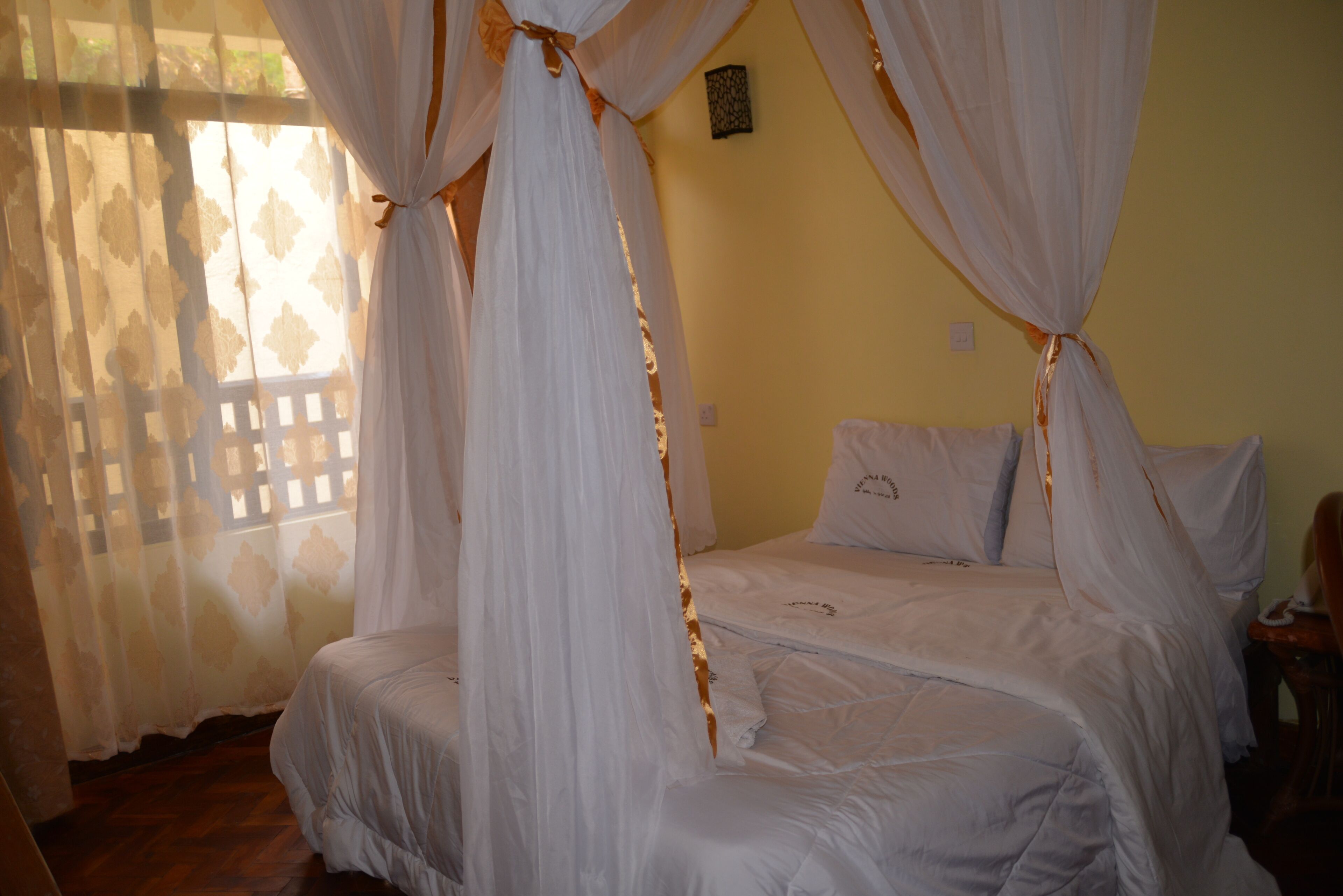 A bedroom with a bed covered by white curtains tied at the corners, a wooden floor, and a small table with a chair.