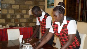 Two individuals in school uniforms serving food at a table with a red plaid tablecloth. - Vienna Woods Holiday Inn Limited (Nakuru)