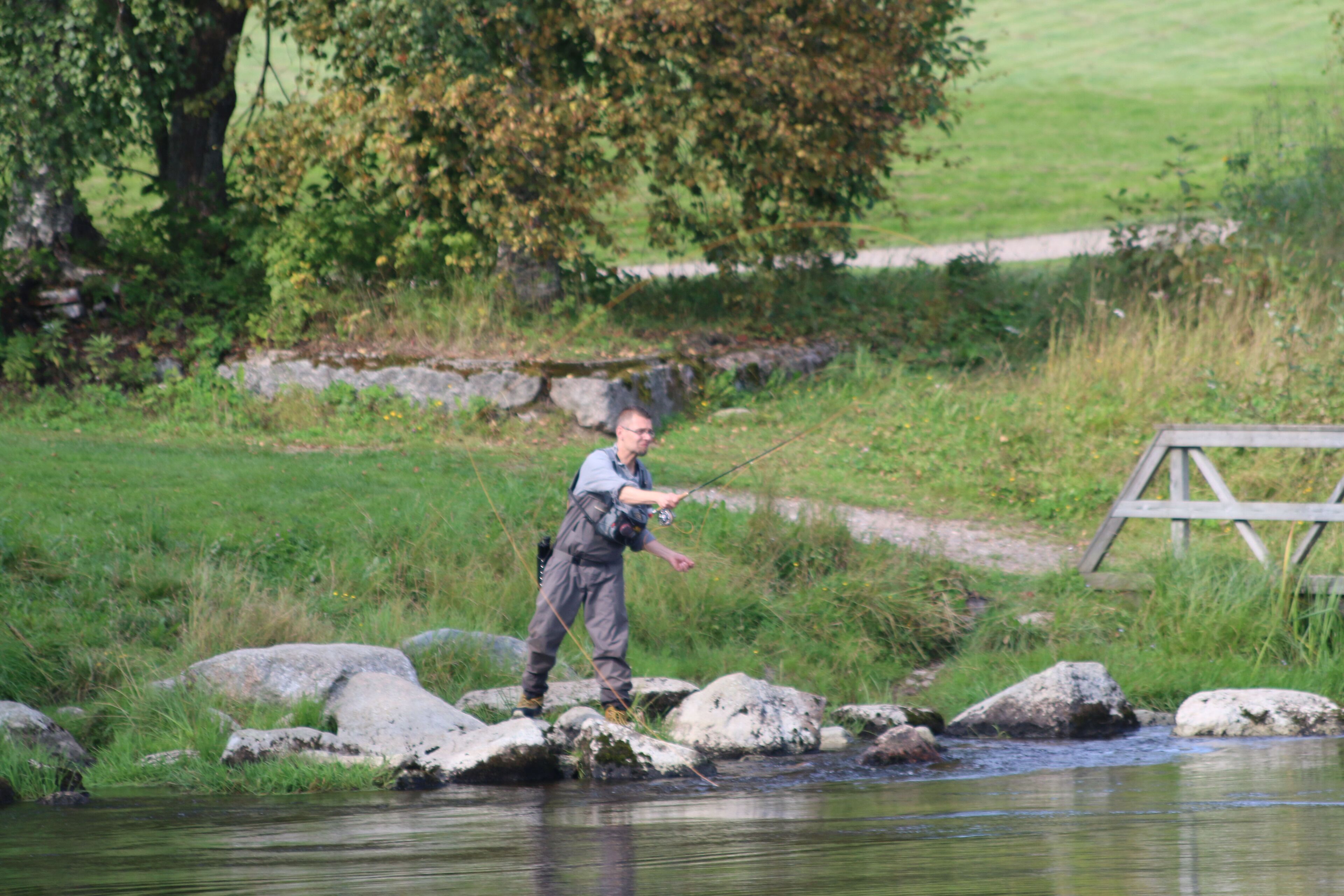 A person fishing by a calm river with rocks and greenery in the background.