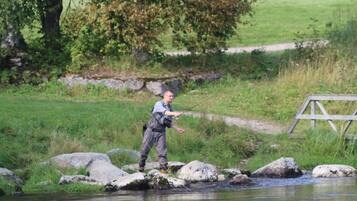 A person fishing by a calm river with rocks and greenery in the background.