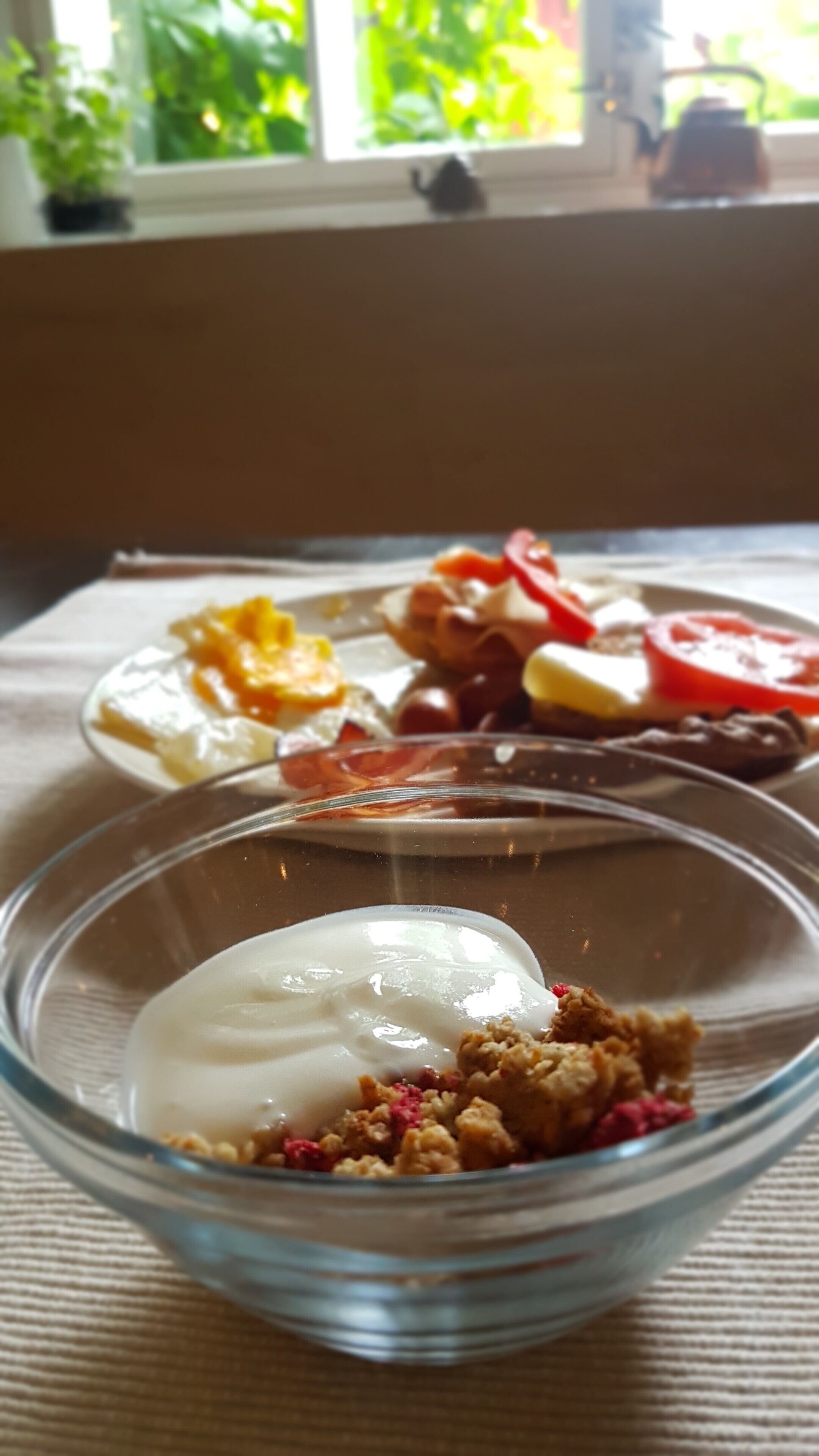 A breakfast setting with a bowl of yogurt and granola, a plate with eggs, tomatoes, and other items.