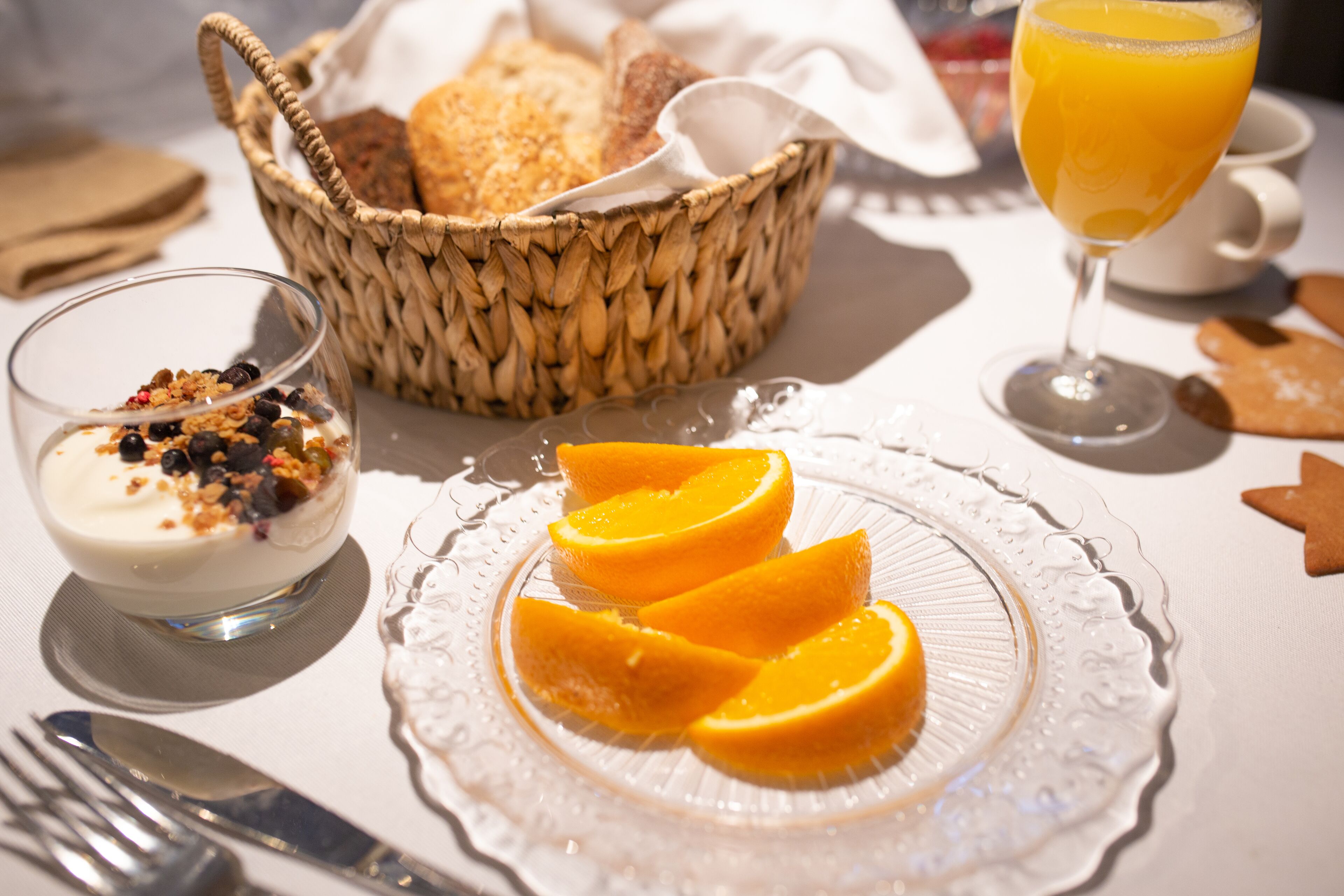 A breakfast setting with a plate of orange slices, a bowl of yogurt with granola, a glass of orange juice, and a basket of bread.