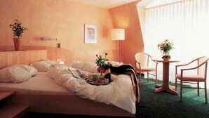 A hotel room with a bed, a bedside table with a lamp, a chair, a small table with a vase, and a window with curtains. - Hotel beim Hirschen (Merzhausen)