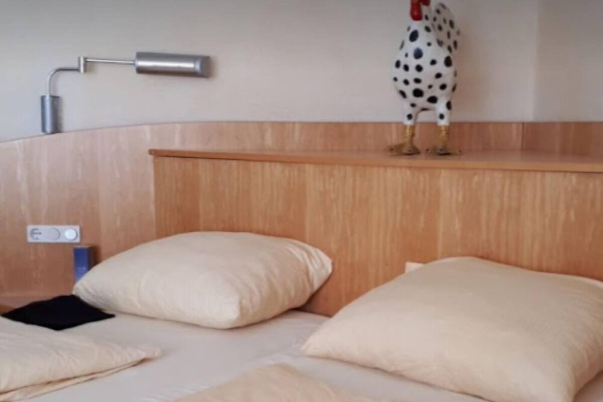 Un lit simple avec une tête de lit en bois, deux oreillers et une petite figurine décorative représentant un coq, placée sur la tête de lit.