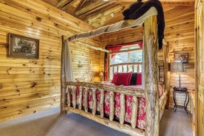 A wooden cabin bedroom with a four-poster bed, a window with red curtains, and a small table with a radio. - Bear Elegance  - Theater Room, Secluded, Heart Shaped Tub!!! (Sevierville)