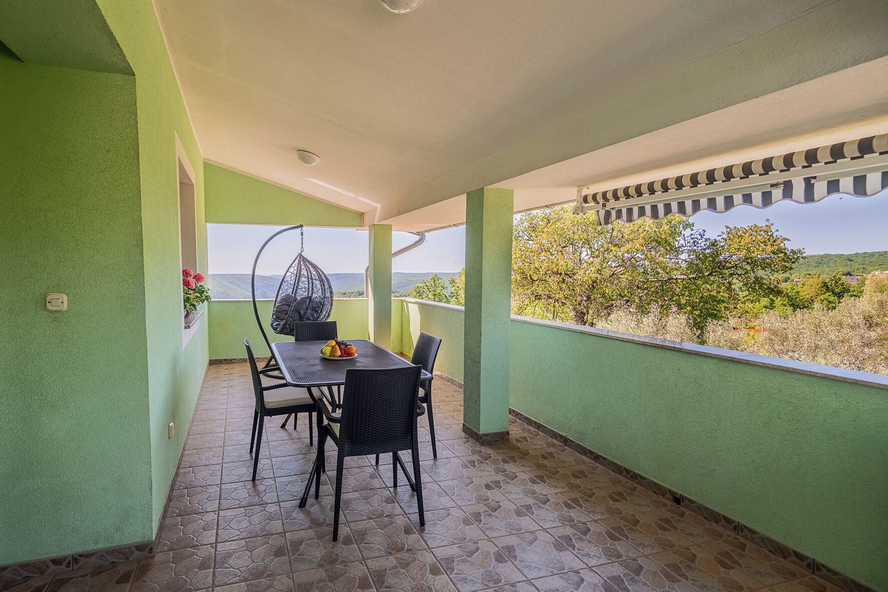 A covered balcony with a table and chairs, a hanging chair, and a view of the landscape.