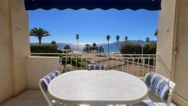 Un balcon avec une table blanche et des coussins à rayures, donnant sur une plage bordée de palmiers et surmontée de montagnes au loin.