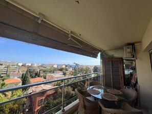 Un balcon doté d’une table en verre et de chaises en osier, offrant une vue sur un quartier résidentiel avec des maisons aux toits rouges et des palmiers.