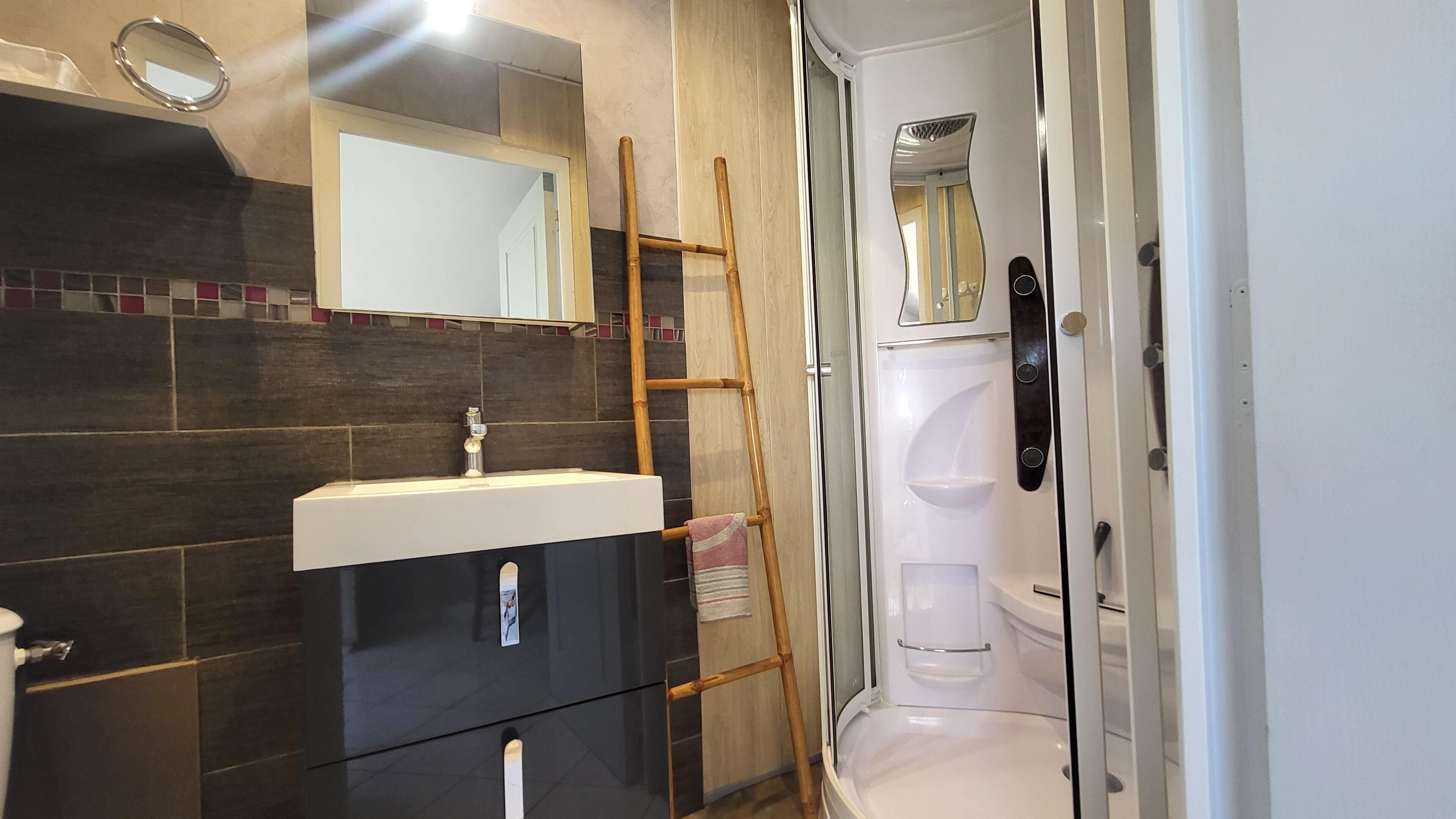 Casa de banho com design moderno, apresentando uma parede revestida de azulejos escuros, uma pia branca, um espelho e uma área de chuveiro com porta de vidro.