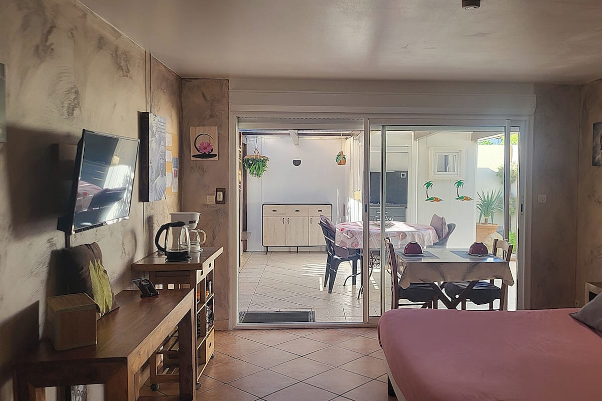 Une chambre avec un lit, un bureau, une chaise et un téléviseur. Une porte coulissante donne sur un balcon équipé d’une table et de chaises.