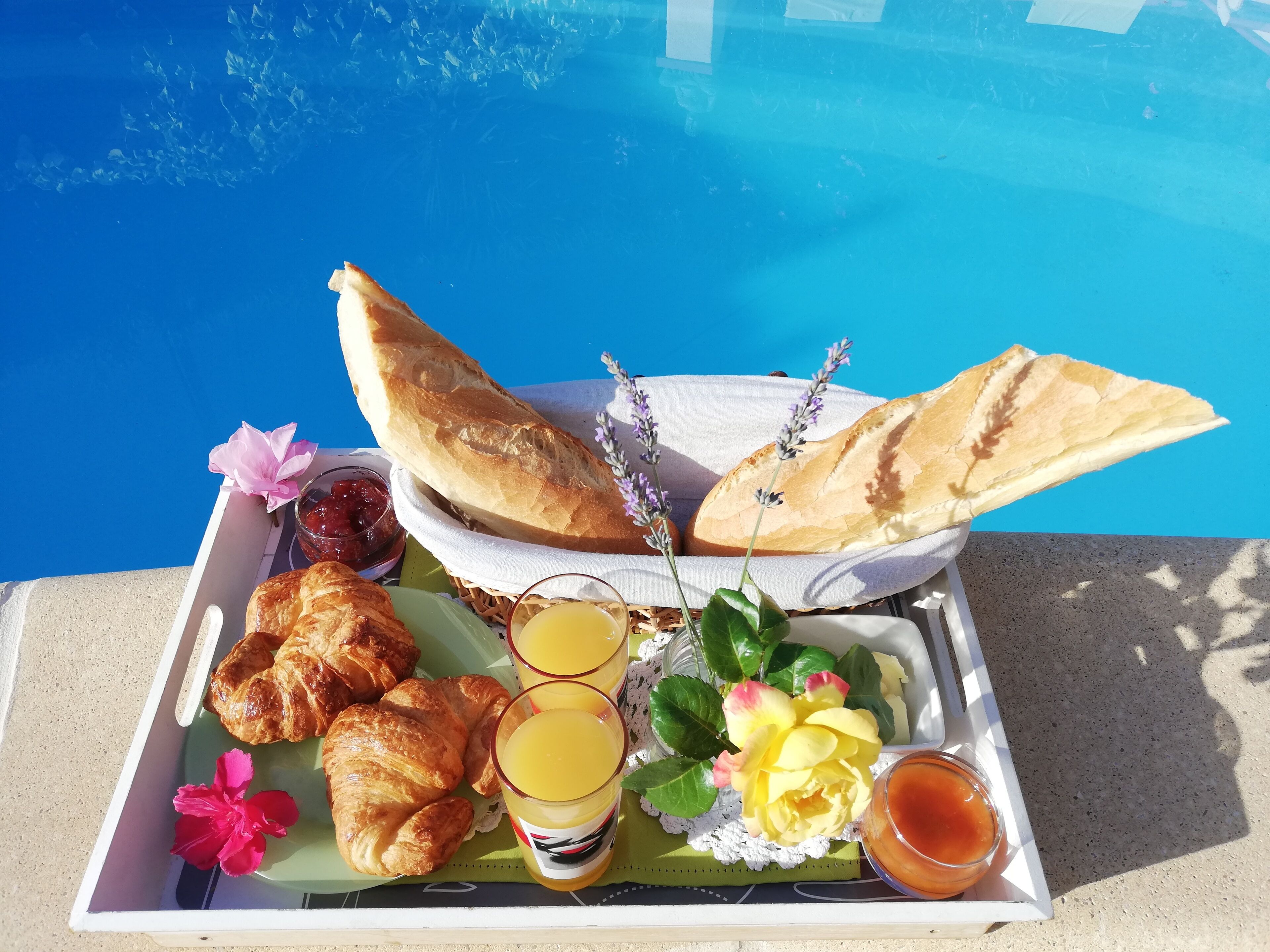 Een dienblad met croissants, brood, sap en bloemen bij een zwembad.
